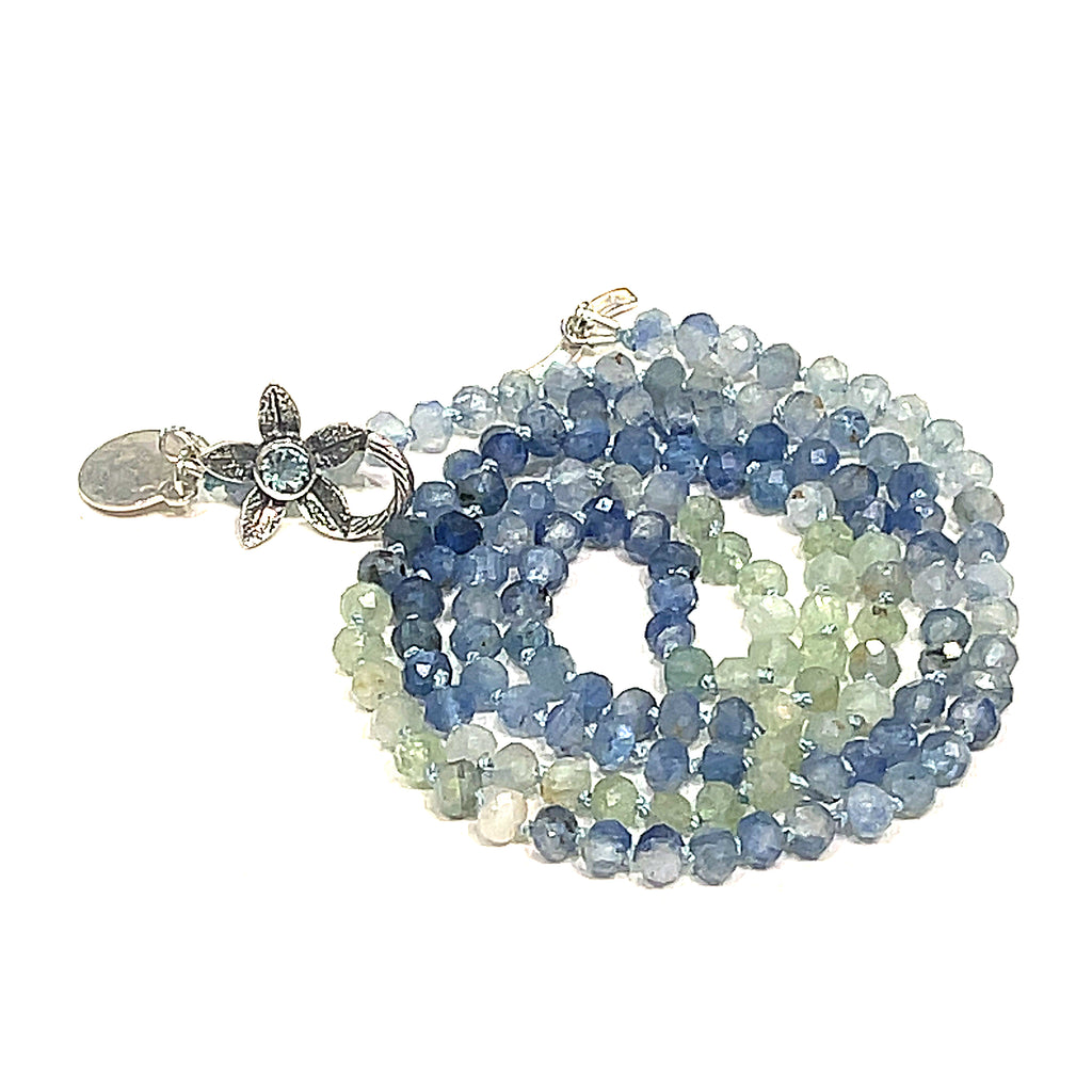 A knotted necklace featuring faceted roundel blue and green kyanite gemstone beads, with a sterling silver flower and a blue topaz gemstone toggle on a white background, front spiral center overview. 