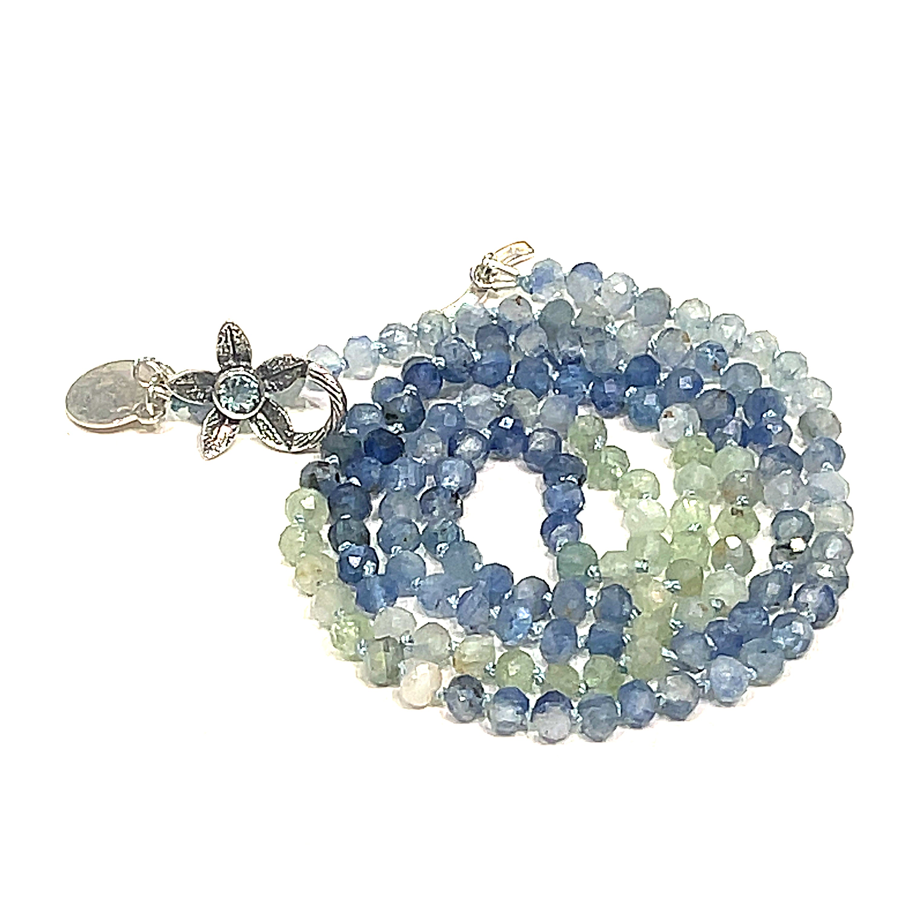 A knotted necklace featuring faceted roundel blue and green kyanite gemstone beads, with a sterling silver flower and a blue topaz gemstone toggle on a white background, front spiral center overview. 