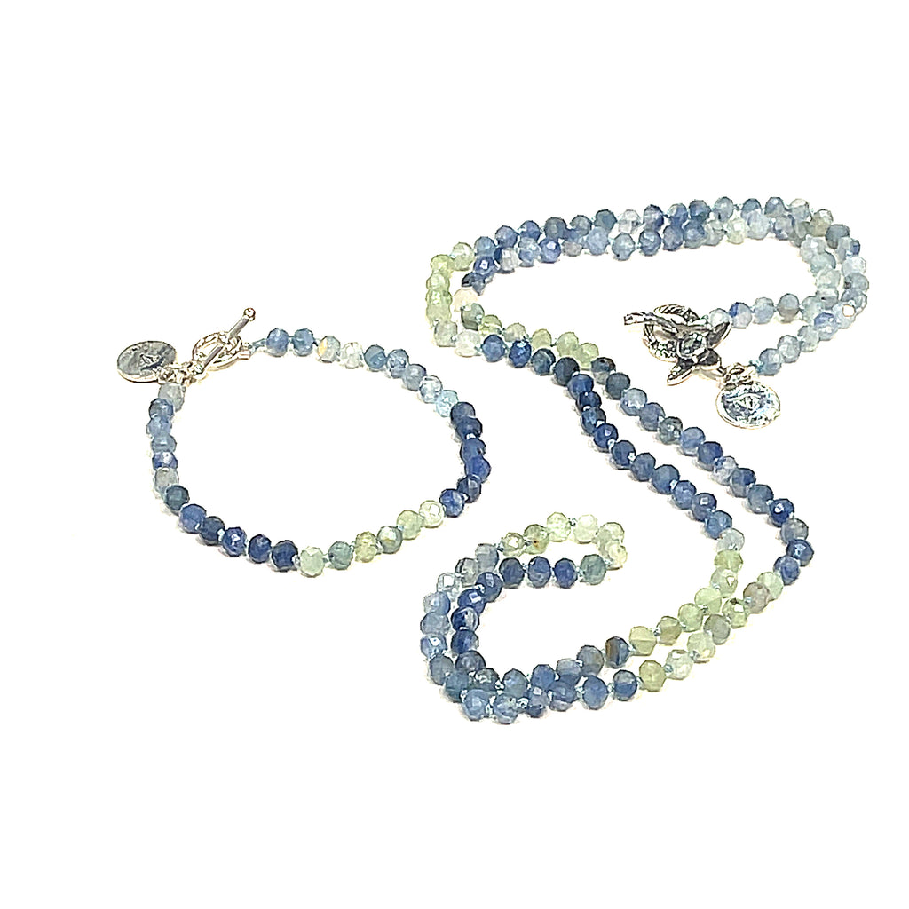 A knotted necklace featuring faceted roundel blue and green kyanite gemstone beads, with a sterling silver flower and a blue topaz gemstone toggle with its matching braclet on a white background, front center, long overview. 