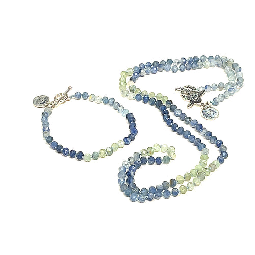 A knotted necklace featuring faceted roundel blue and green kyanite gemstone beads, with a sterling silver flower and a blue topaz gemstone toggle with its matching braclet on a white background, front center, long overview. 