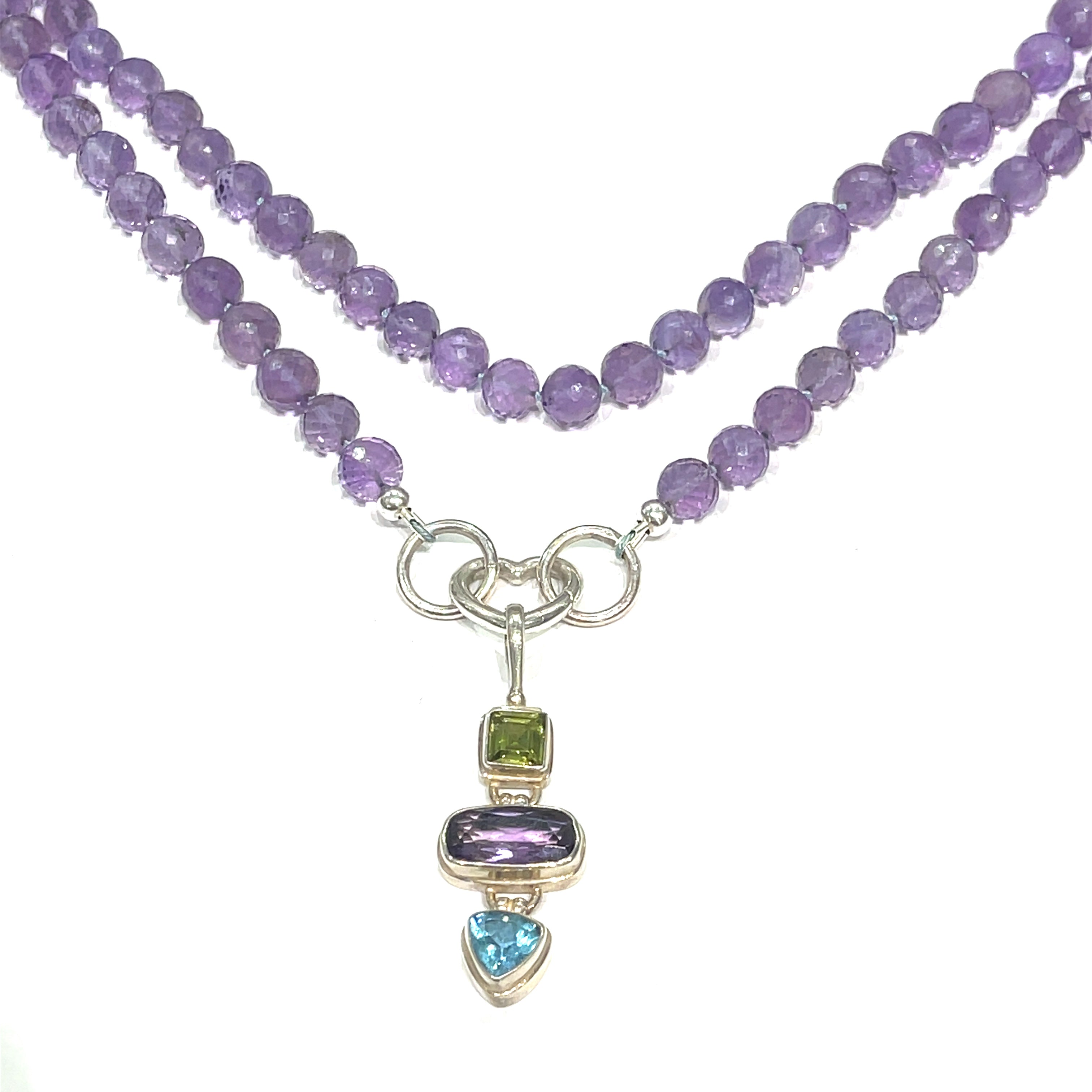 A knotted necklace featuring round Amethyst gemstones, a sterling silver heart clasp, and a silver gemstone pendant with Peridot and Blue Topaz, on a white background, long hanging center front overview. 