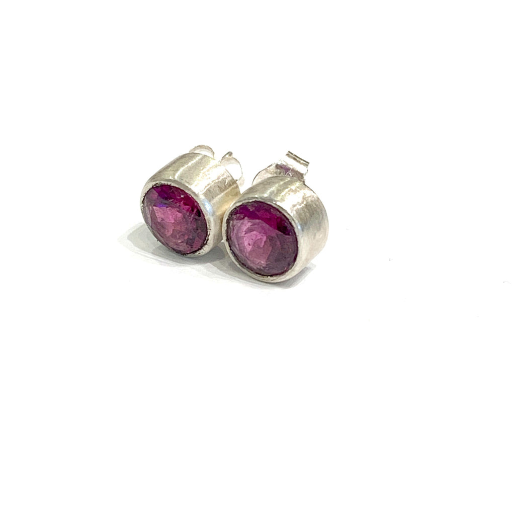 A pair of faceted ruby gemstone round tube-set earrings with sterling silver tube settings on a white background, front left angle overview.