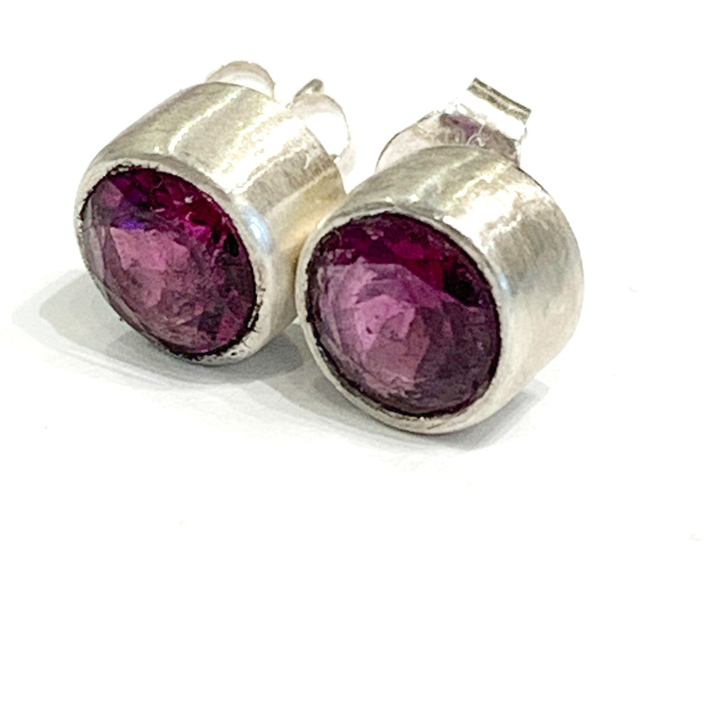 A pair of faceted ruby gemstone round tube-set earrings with sterling silver tube settings on a white background, front left angle view.