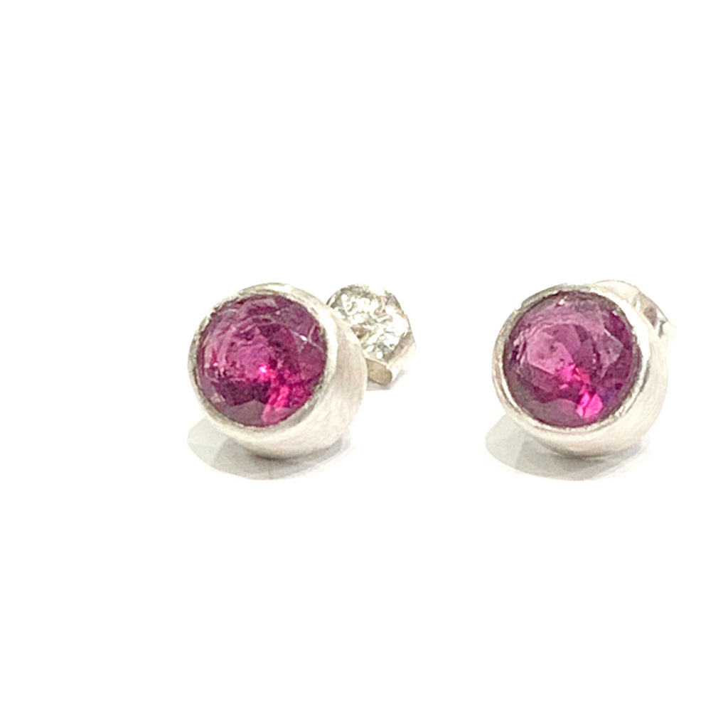 A pair of faceted ruby gemstone round tube-set earrings with sterling silver tube settings on a white background, top center close up left angle overview.