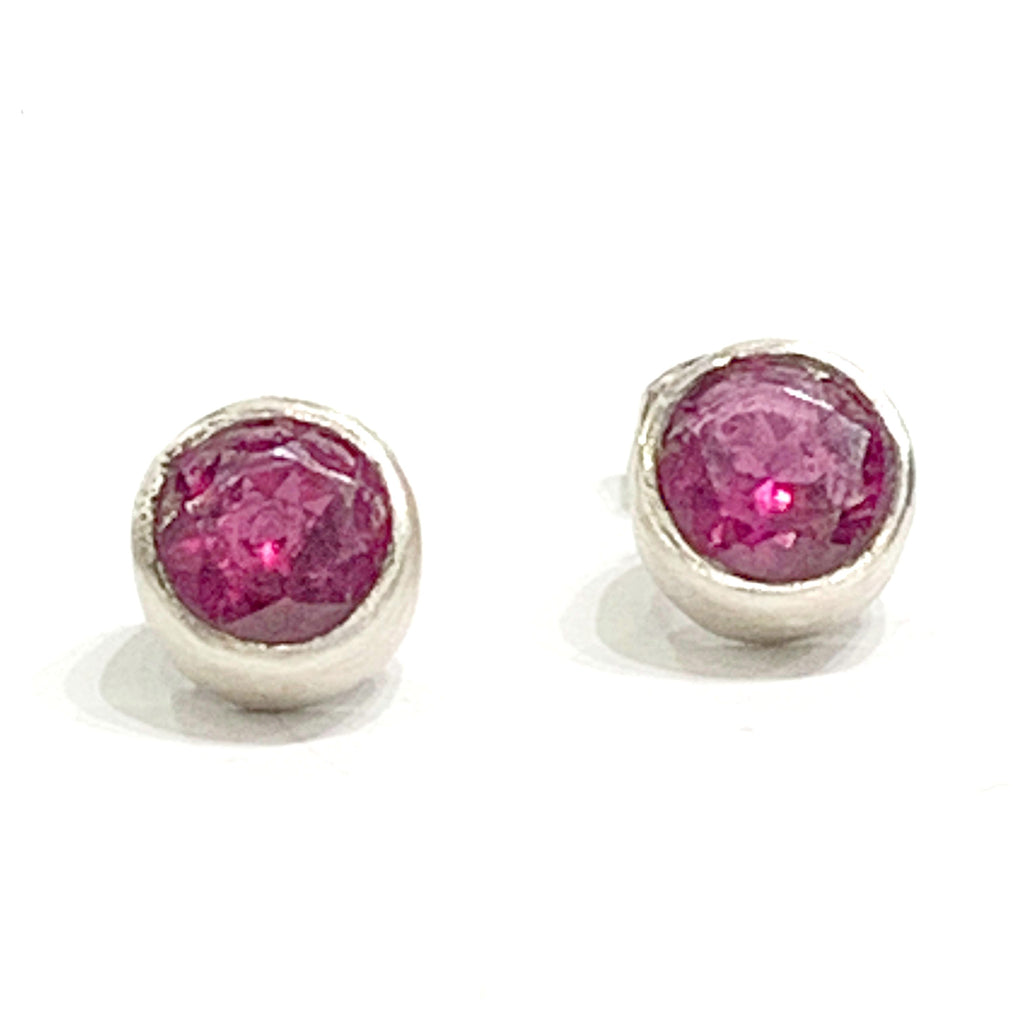 A pair of faceted ruby gemstone round tube-set earrings with sterling silver tube settings on a white background, top center close up overview.