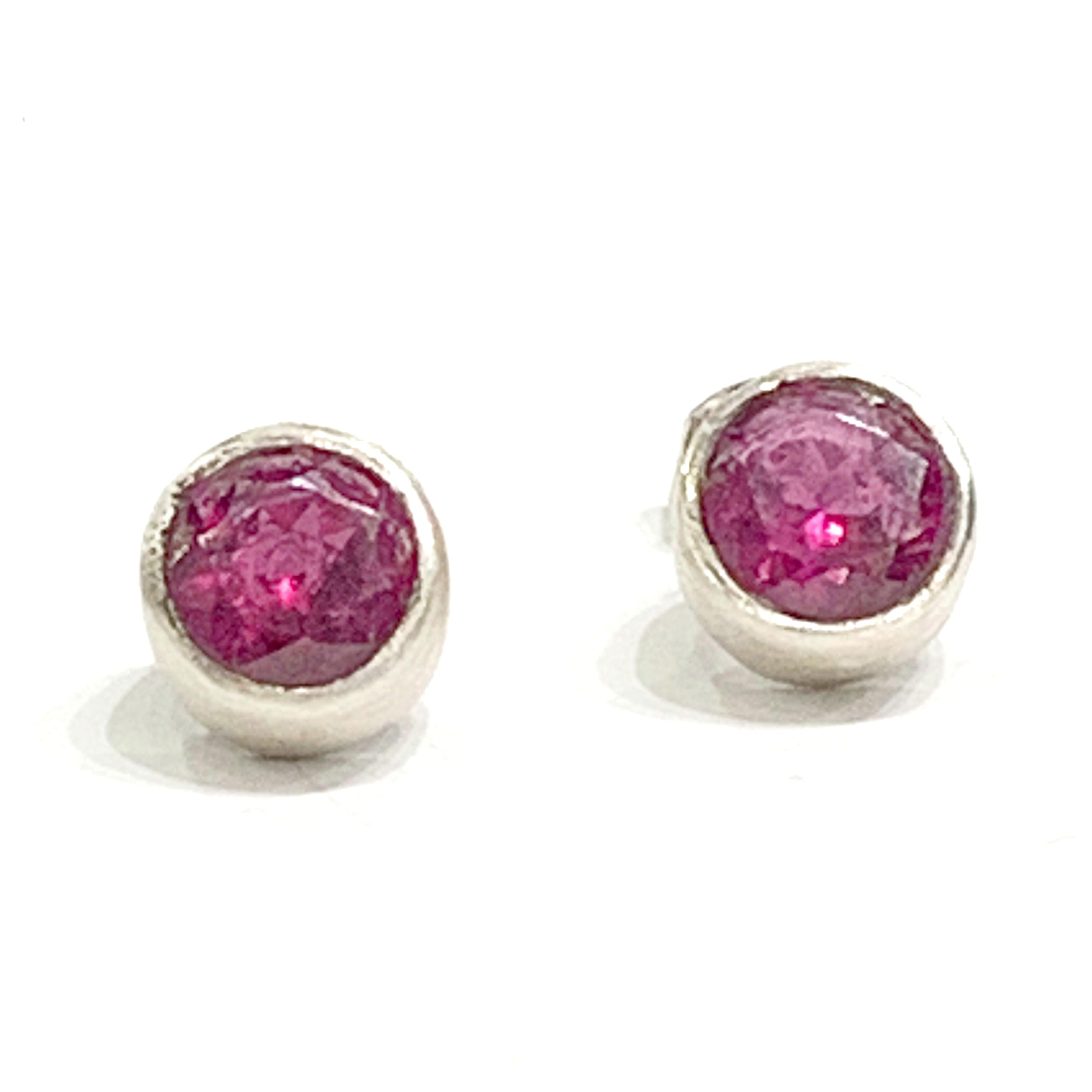 A pair of faceted ruby gemstone round tube-set earrings with sterling silver tube settings on a white background, top center close up overview.