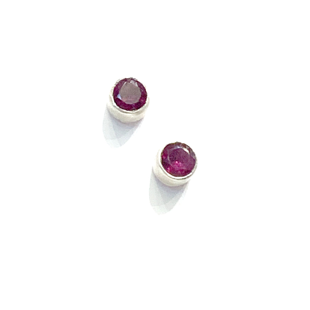 A pair of faceted ruby gemstone round tube-set earrings with sterling silver tube settings on a white background, top diaganol view.