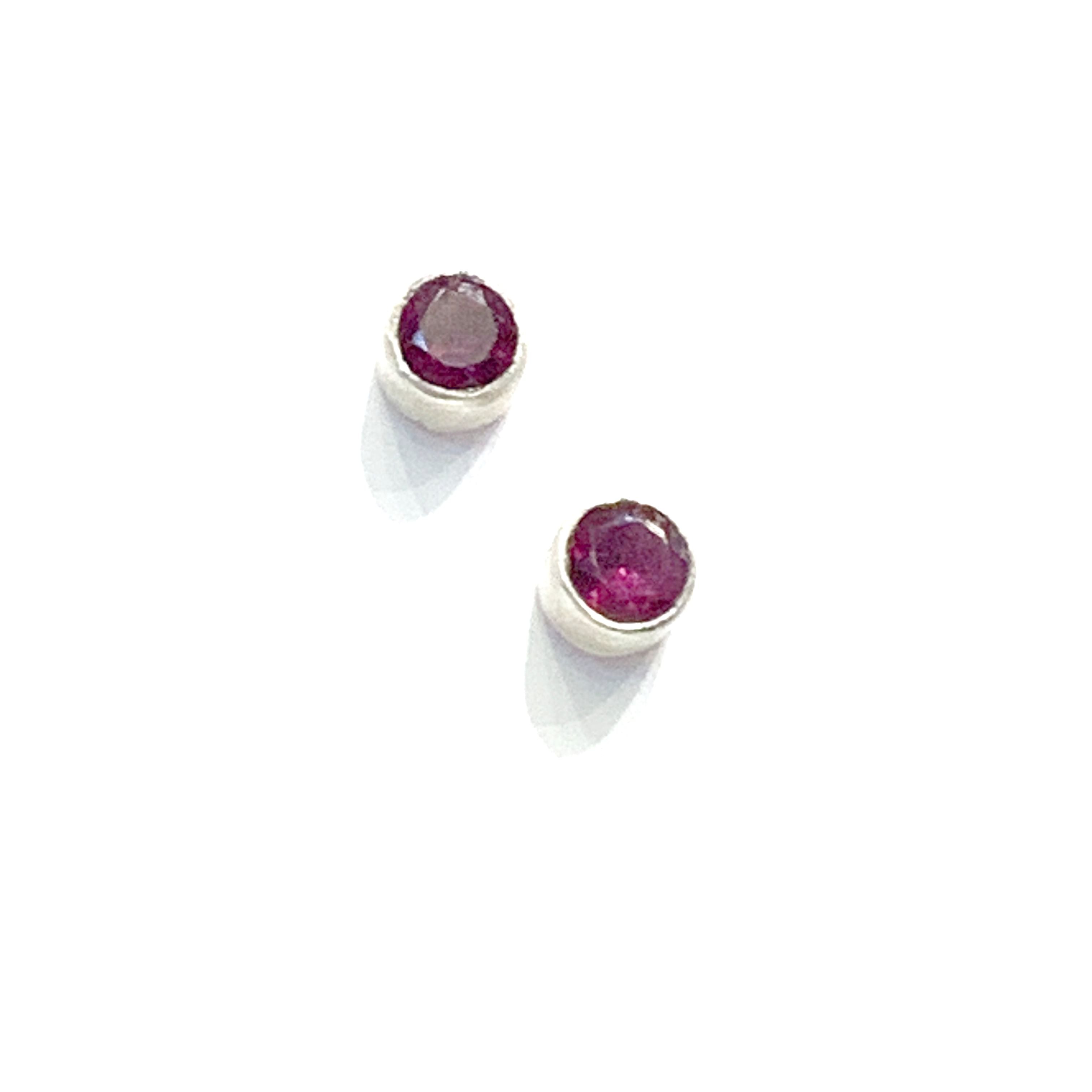 A pair of faceted ruby gemstone round tube-set earrings with sterling silver tube settings on a white background, top diaganol view.