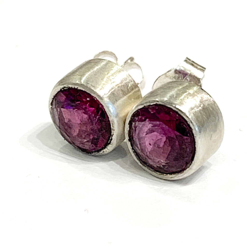 A pair of faceted ruby gemstone round tube-set earrings with sterling silver wire basket settings on a white background, front-left angle view.
