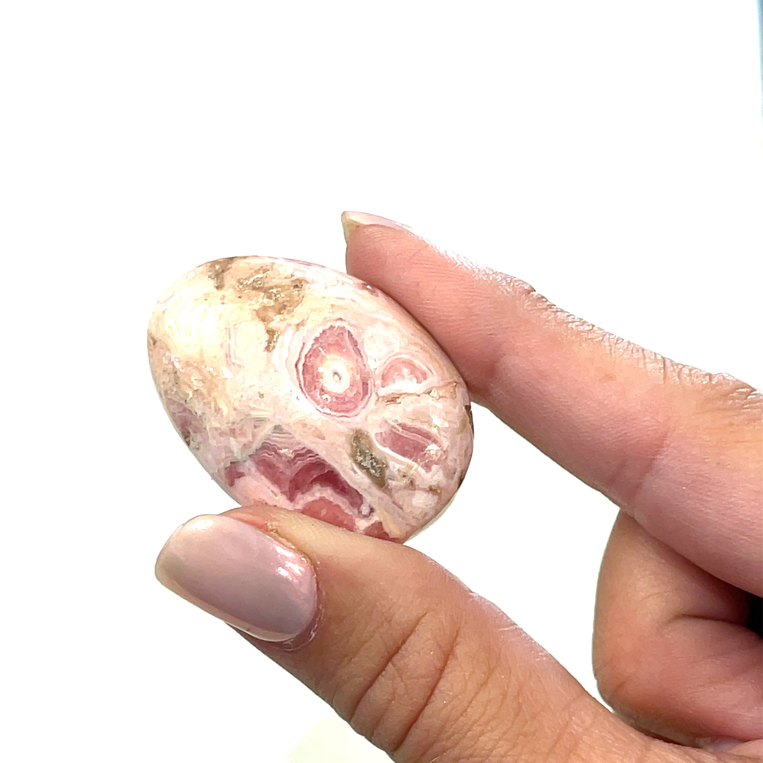 A polished Rhodochrosite gemstone palm stones with varying shades of pink to pale brown colors and white streaks on a white background front view in a models finger tips. 