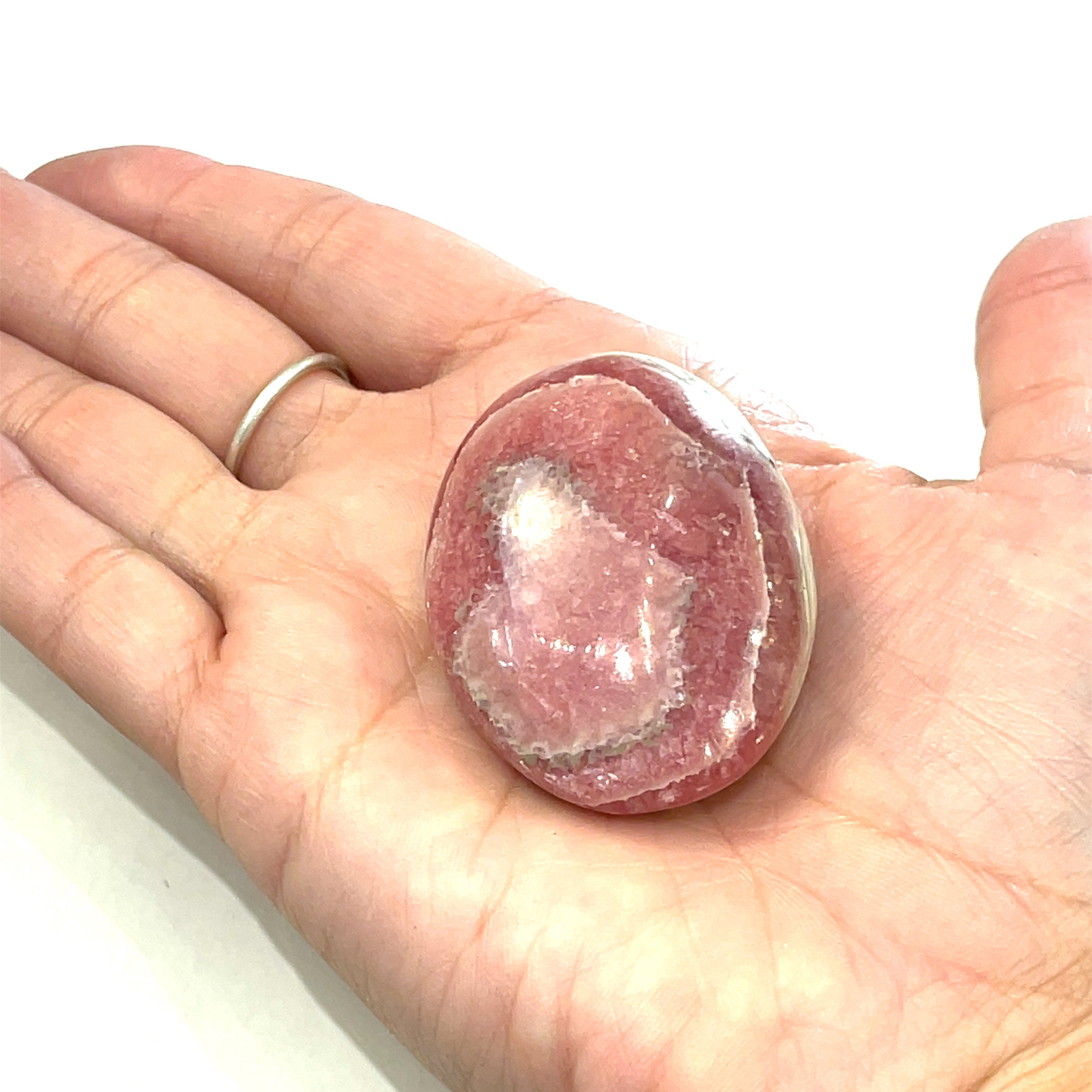 A polished Rhodochrosite gemstone palm stones with varying shades of pink to pale brown colors and white streaks on a white background front view in a models hand. 
