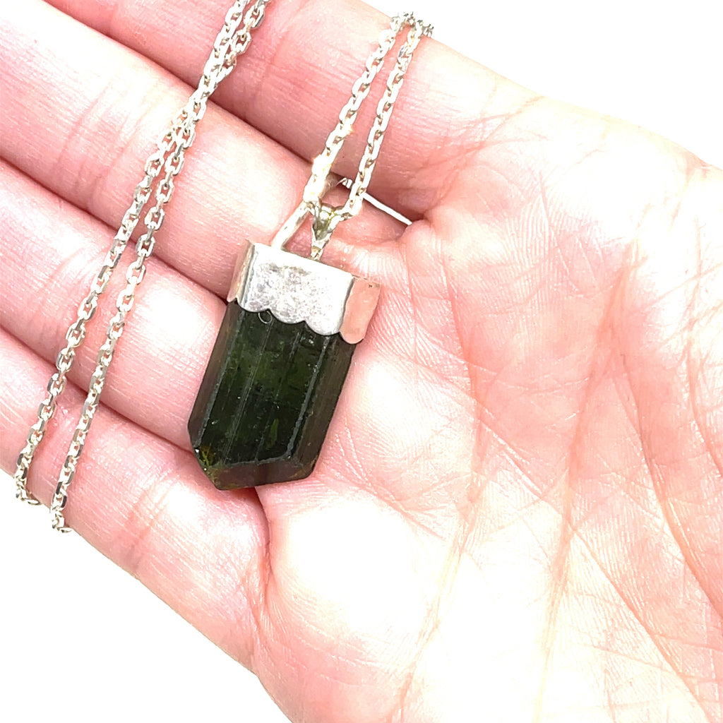 A raw Green Tourmaline pendant in a models hand on a white background center hanging overview. 