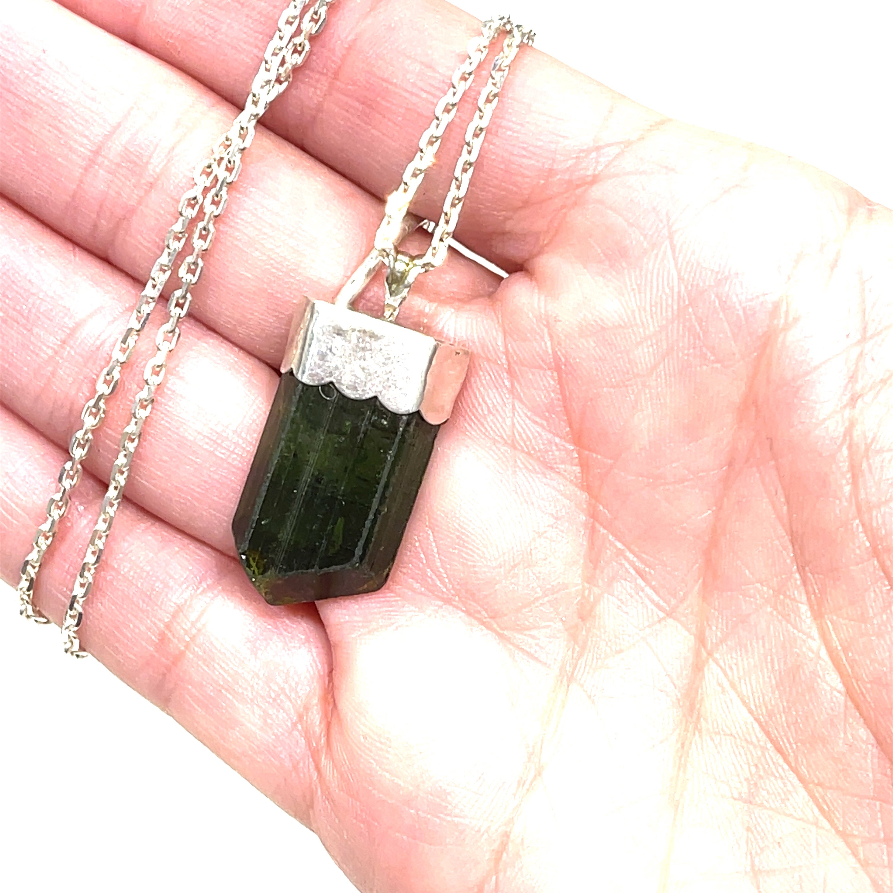 A raw Green Tourmaline pendant in a models hand on a white background center hanging overview. 