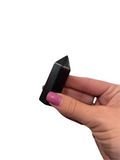 A small sungite gemstone tower in a models finger tips dispalyed on a white background, front view. 