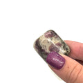 A tumbled Eudialyte gemstones with a purple and gray color palette on a white background, front overview. in a models hand.