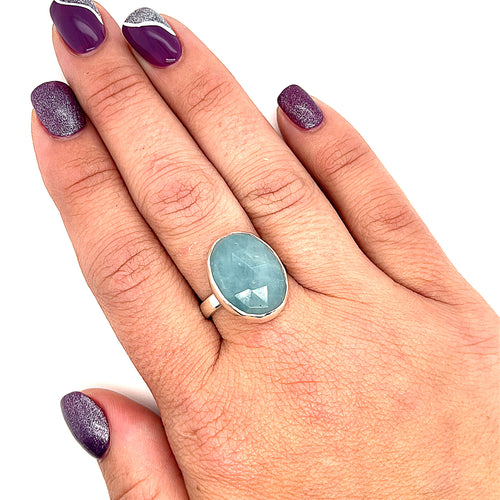 Aquamarine gemstone ring with a bezel-set design, worn on a model's finger, with a matching nail polish in the background front overview.