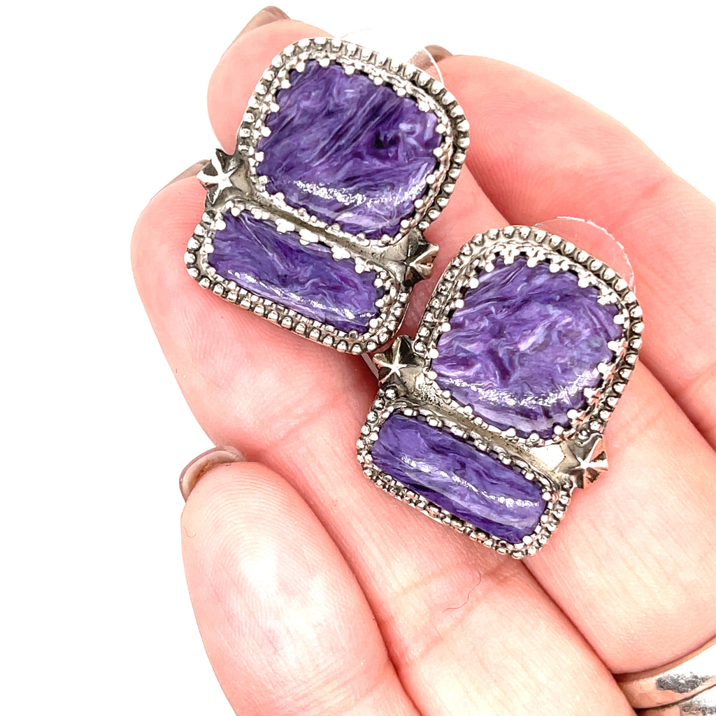 Rectangle Charoite double stone sterling silver earrings with swirling purple patterns studs in a models hand to show an up-close front view on a white background.