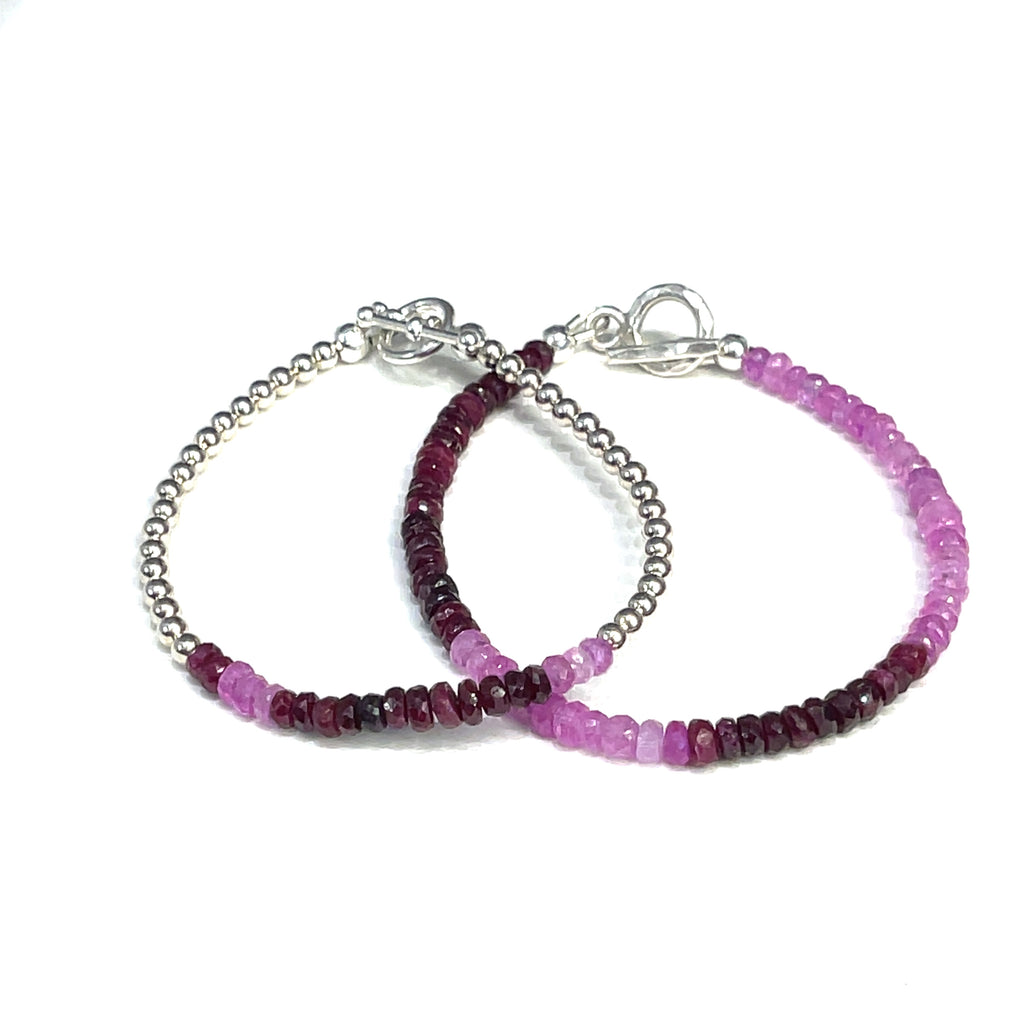 Two genuine ruby gemstone beaded bracelets with gold-filled toggle clasps, featuring faceted roundel rubies ranging in color from purple to red, on a white background front overview.