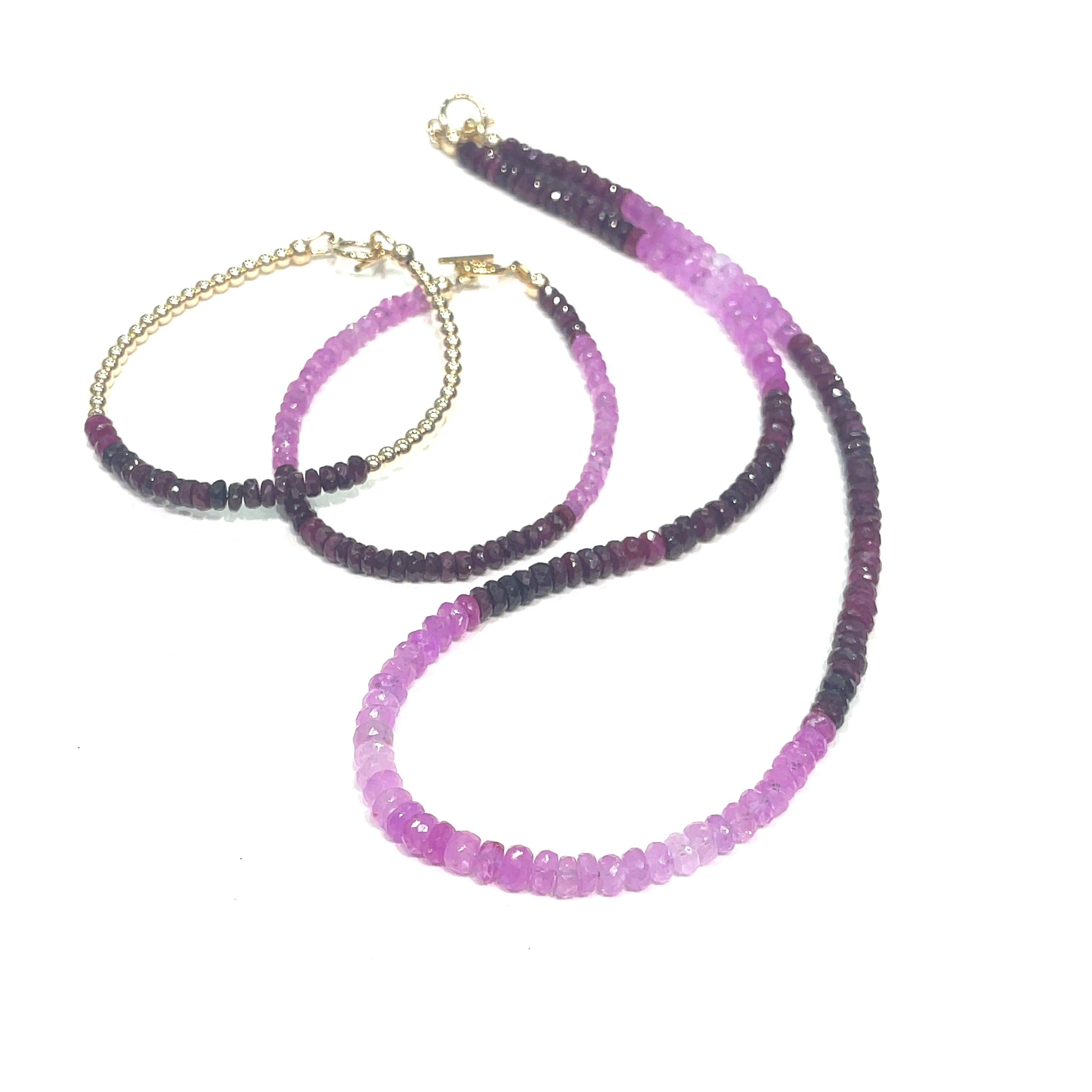 Two genuine ruby gemstone beaded bracelets with gold-filled toggle clasps, featuring faceted roundel rubies ranging in color from purple to red, on a white background front overview with its matching necklace.