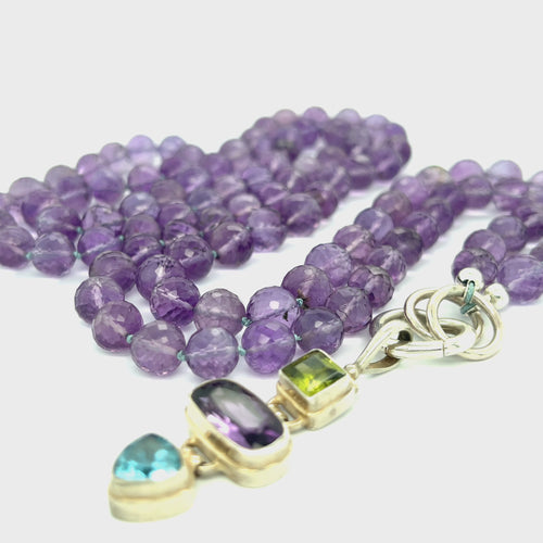 A knotted necklace featuring round Amethyst gemstones, a sterling silver heart clasp, and a silver gemstone pendant with Peridot and Blue Topaz, on a white background, center front overview.  video